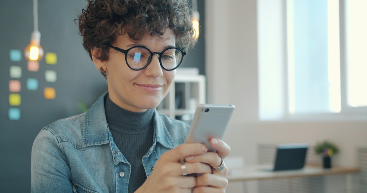 Mulher de óculos sorrindo ao olhar o celular em um escritório